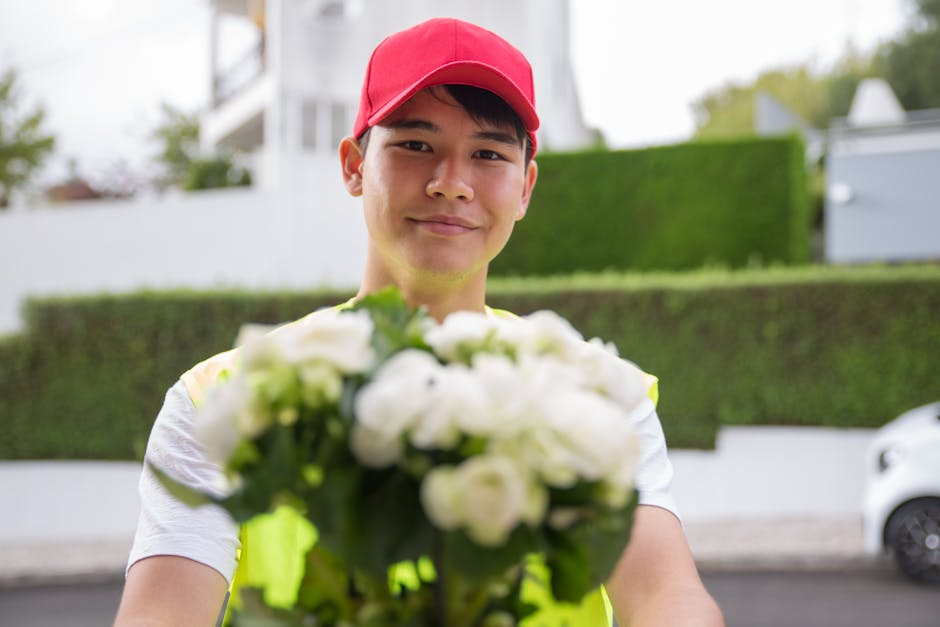 A male flower delivery courier wearing a red cap and high-visibility vest is holding a bouquet of fresh white roses and other white flowers. The bouquet is arranged with green foliage and wrapped in c